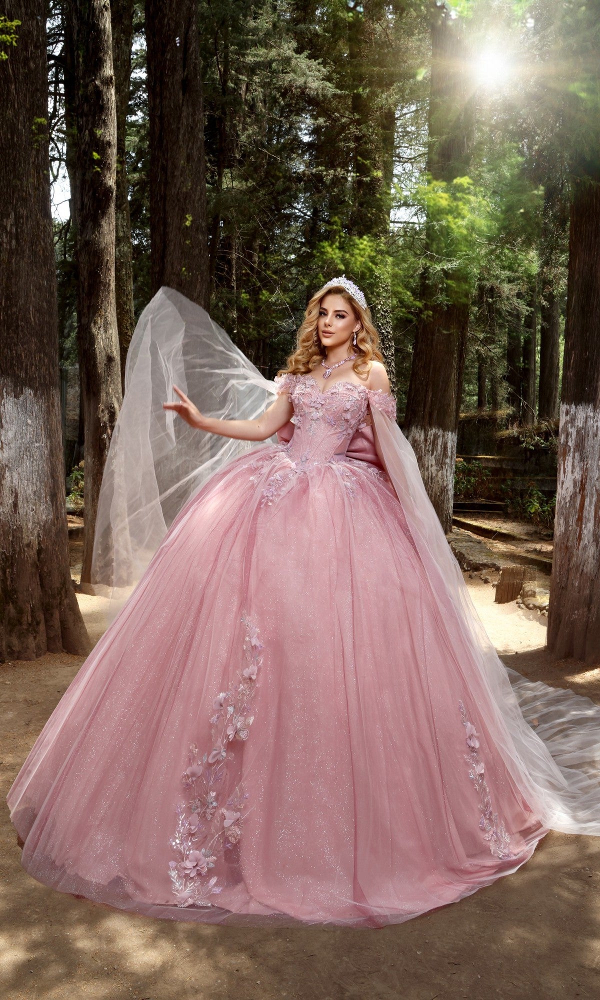A woman poses in a sunlit forest wearing the Quinceanera Dress Q117 By Nina Canacci in Blush/Burgundy, featuring floral details, a glitter tulle skirt, a long veil, and a tiara, with one arm raised.